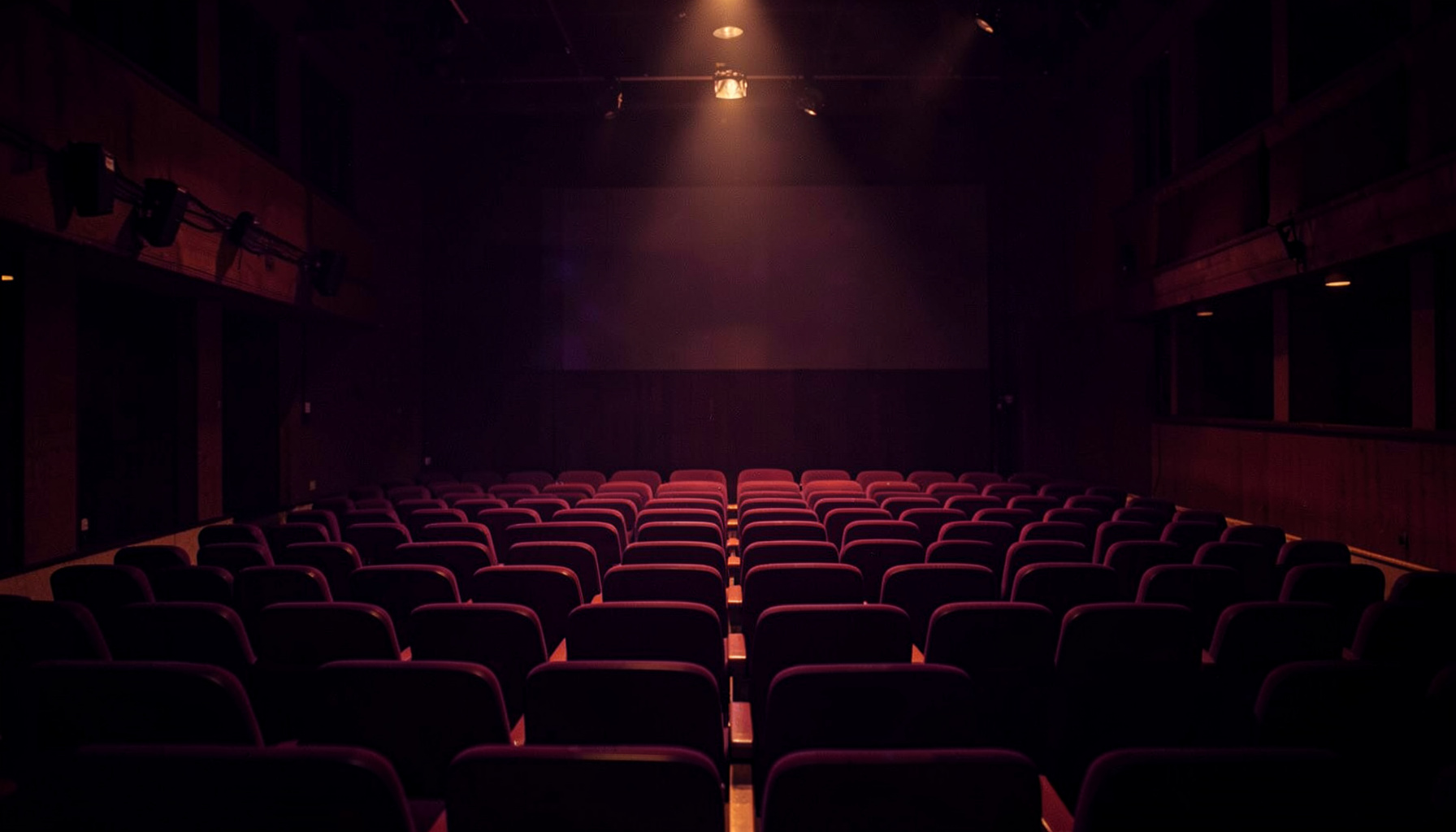 A warm, softly lit theatre stage with deep purple and amber tones, empty seats fading into shadow, evoking anticipation and artistic possibility.